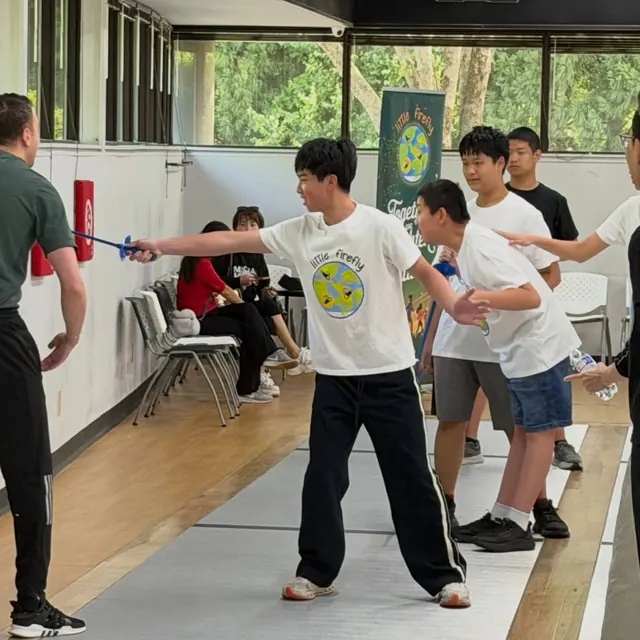 A Little Firefly participant practicing fencing with a coach and volunteers nearby